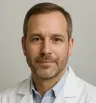 A headshot of a middle-aged man wearing a white lab coat and a light blue collared shirt, with short brown hair and a neutral expression.