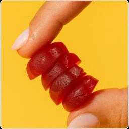 A hand holding a red gummy worm candy between thumb and index finger with white nails against a yellow background