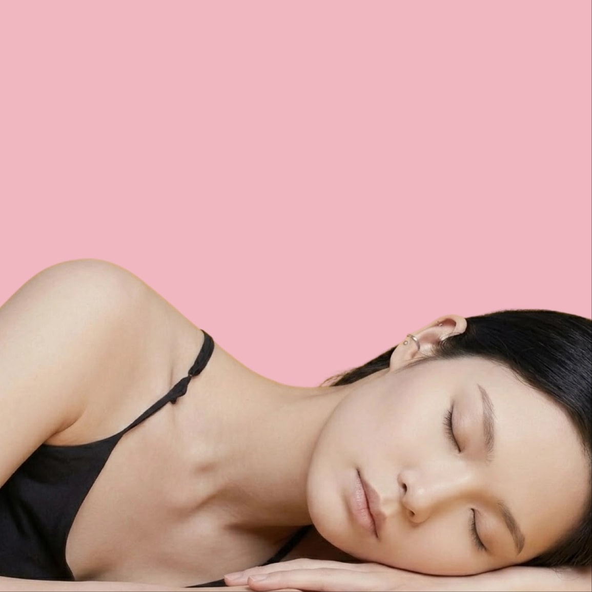 A woman with dark hair and fair skin wearing black lingerie, lying on her side with her head resting on her hand, eyes closed, against a pink background