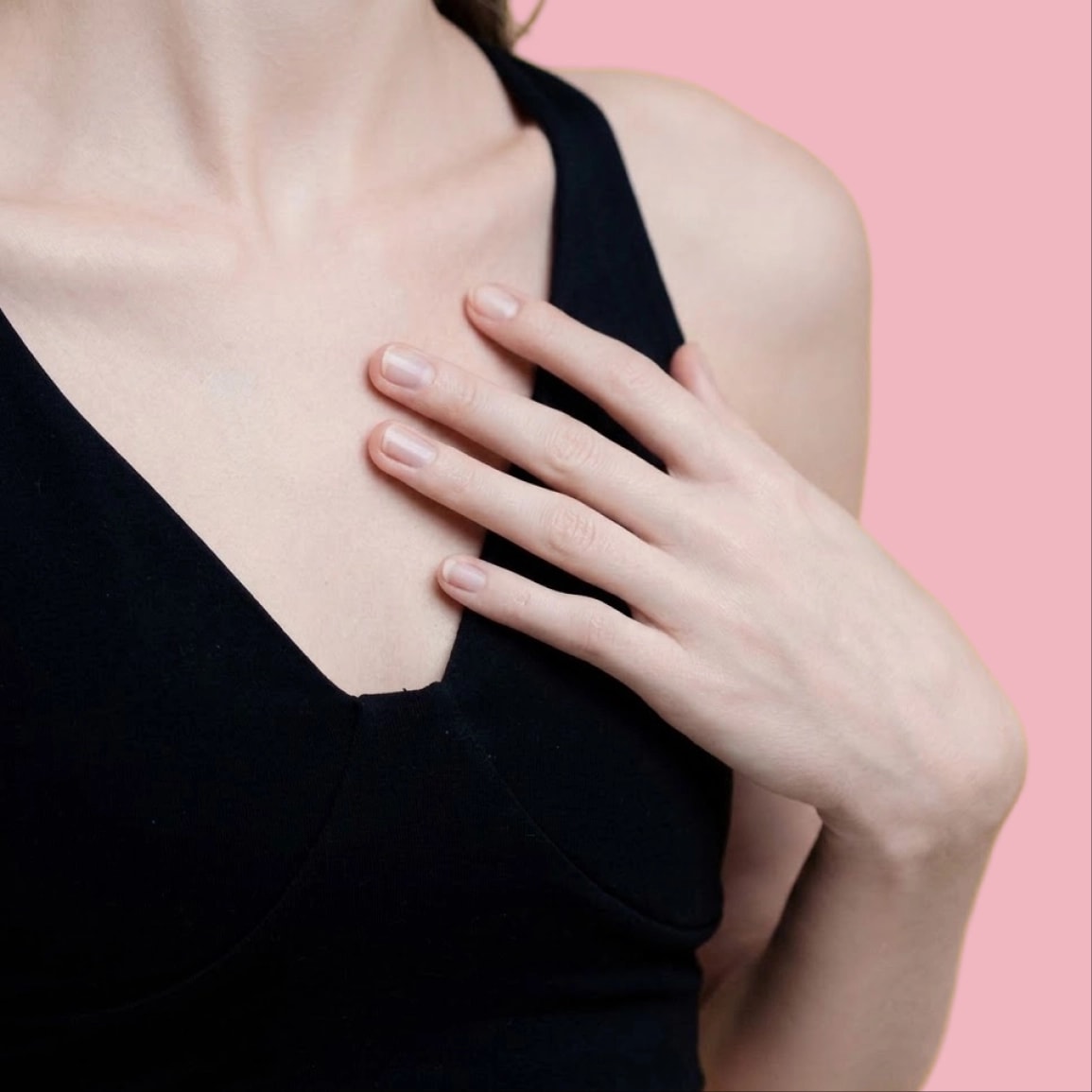 A woman in a black top with her hand on her chest, against a pink background
